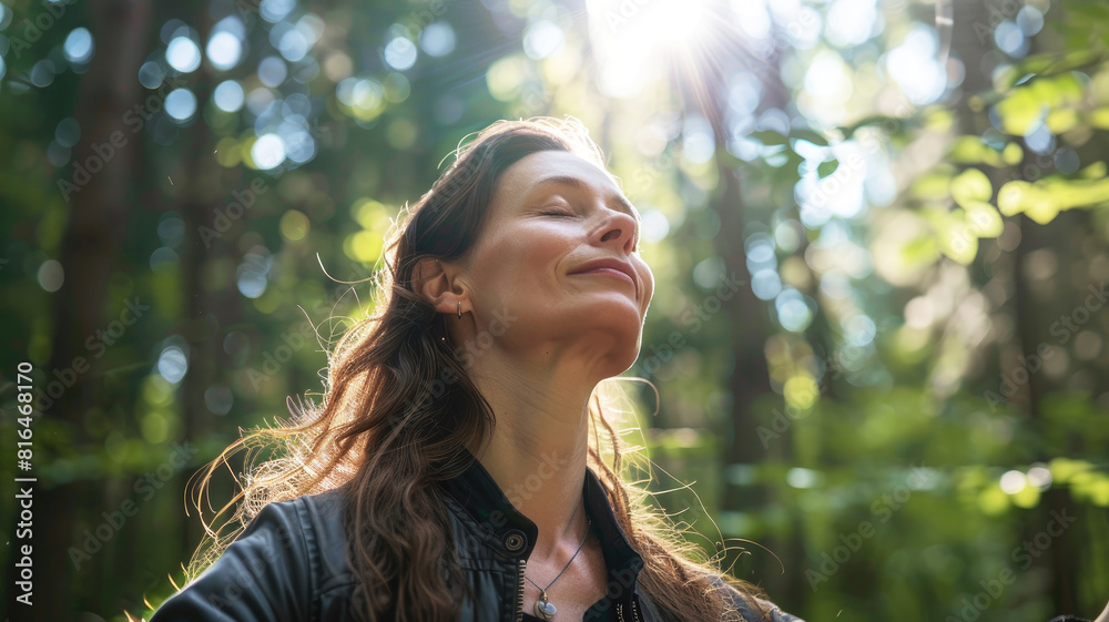 A beautiful and peaceful woman in her thirties with her eyes closed and arms outstretched, breathing the fresh air in the forest with sunlight shining through the trees