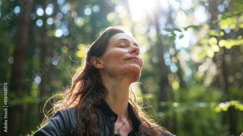 A beautiful and peaceful woman in her thirties with her eyes closed and arms outstretched  breathing the fresh air in the forest with sunlight shining through the trees