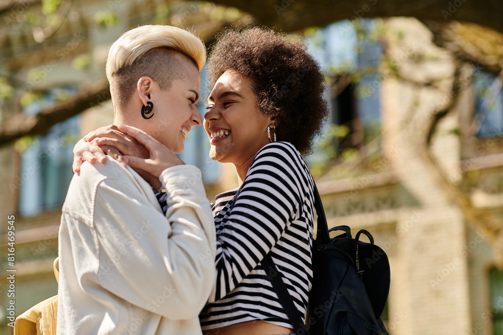 Two young women, one with dark hair and the other with blonde hair, hugging tightly in front of a lush green tree.