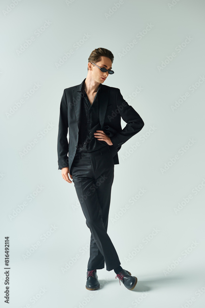 A young queer person in a sleek black suit striking a confident pose in a studio against a grey background. A young queer person in a sleek black suit striking a confident pose in a studio against a grey background.