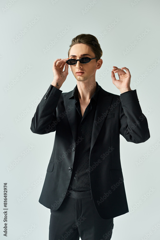 A young queer person strikes a confident pose in a black suit and sunglasses against a grey background. A young queer person strikes a confident pose in a black suit and sunglasses against a grey background.