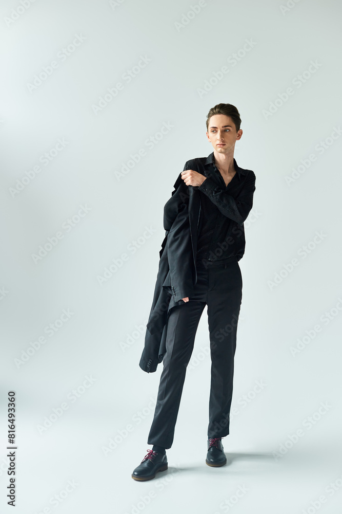 A young queer person, exuding pride, strikes a pose in a studio wearing a black coat and pants on a grey background. A young queer person, exuding pride, strikes a pose in a studio wearing a black coat and pants on a grey background.