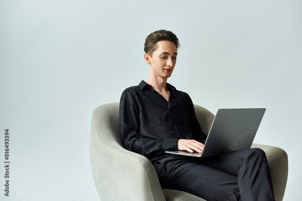 A young queer person, proudly expressing their identity, sitting in a chair using a laptop in a studio with a grey background. A young queer person, proudly expressing their identity, sitting in a chair using a laptop in a studio with a grey background.