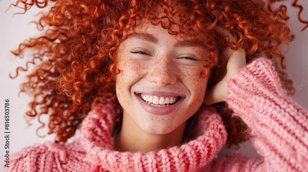 A woman with red curls smiles, wearing a pink sweater as she touches her ear A woman with red curls smiles, wearing a pink sweater as she touches her ear