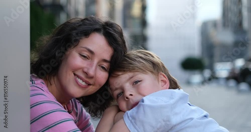 Cute caucasian brunette mother and kid son sitting outdoors face to face talking to each other. Motherhood or family day concept. 8 years old boy with mother in bright clothes enjoying summer day