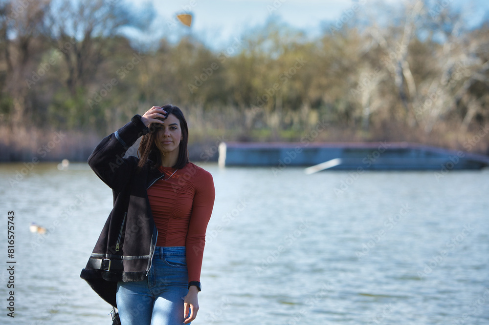 Young, beautiful brunette woman, with short hair and jeans and orange shirt with jacket over her shoulder, walks through the forest. In the background a lake. Young, beautiful brunette woman, with short hair and jeans and orange shirt with jacket over her shoulder, walks through the forest. In the background a lake.