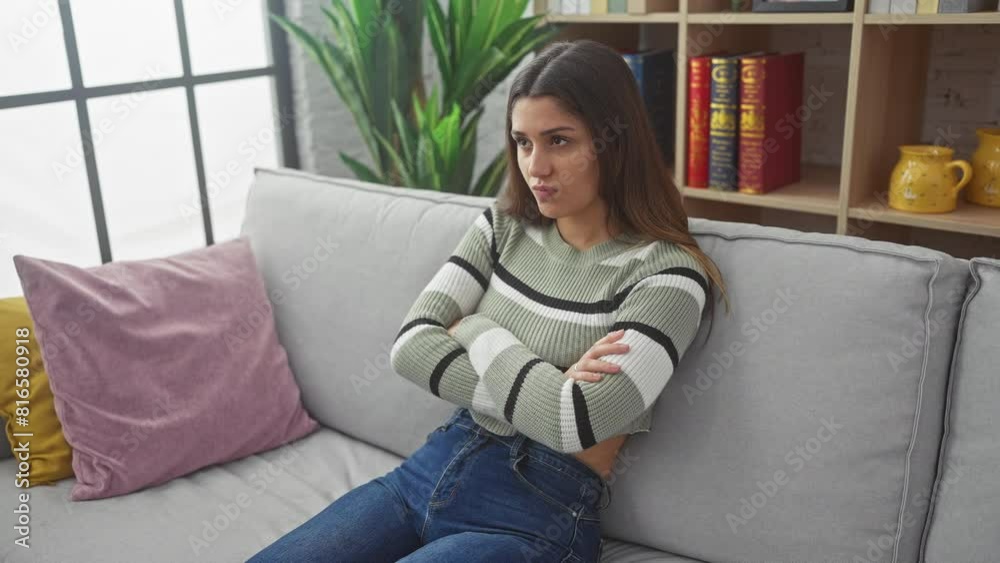 Distressed young hispanic woman at home, a nervous doubter, sitting on the sofa, wearing a sweater, with a serious frown, negative expression and crossed arms