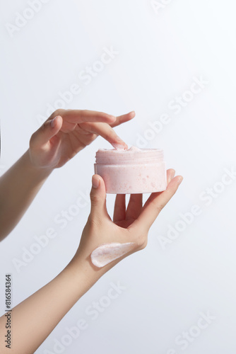 A jar of pink exfoliating scrubs hold by a beautiful hand with a pink smudge on, the other hand is touching cream in the jar. Photo was taken on white background from front view, copy space © Tuan  Nguyen 