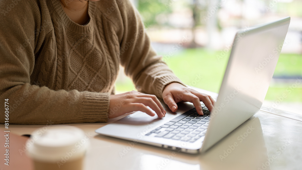 Fototapeta premium A woman in a cozy knitted sweater working on her computer, typing on the laptop keyboard.