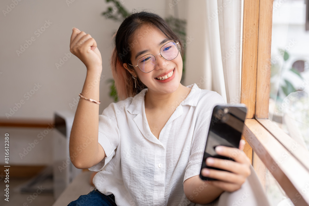 A young, cheerful Asian woman celebrating good news on her smartphone indoors. A young, cheerful Asian woman celebrating good news on her smartphone indoors.
