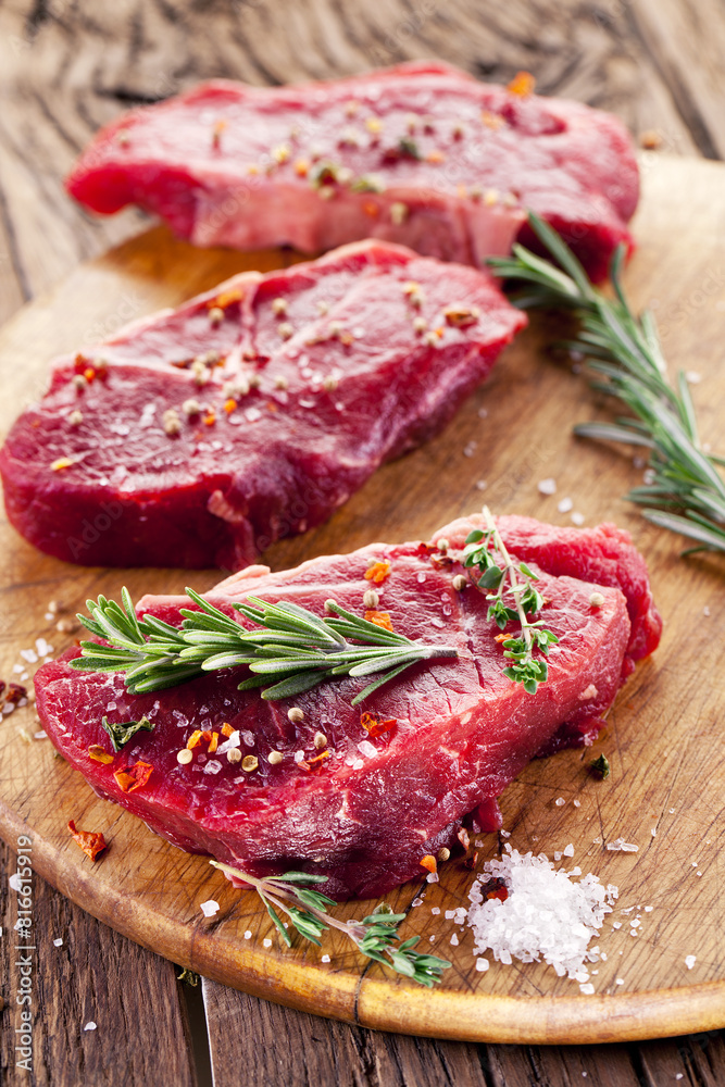 Raw beef steaks with herbs and spices ready for cooking are on wooden board.