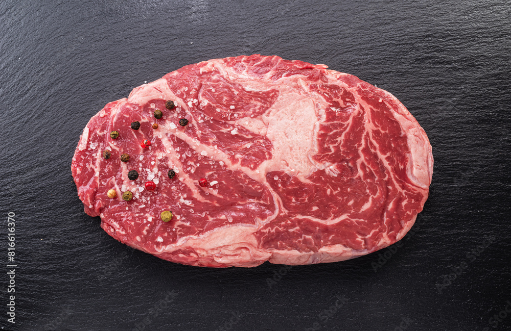 Raw ribeye steak with pepper corns and rosemary on graphite serving board, top view. Raw ribeye steak with pepper corns and rosemary on graphite serving board, top view.