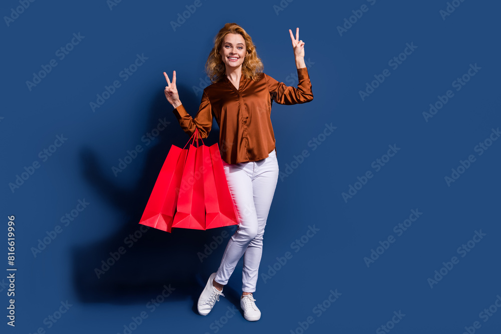 Fototapeta premium Full length photo of adorable woman dressed silk shirt hold shopping bags showing v-sign symbol isolated on dark blue color background