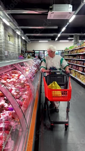 a woman with a cart in a supermarket selects food products from a display case with meat. old woman, vertical video