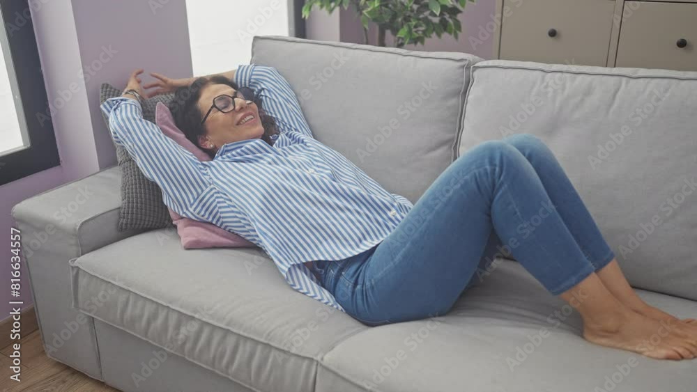 A relaxed middle-aged woman stretching on a couch indoors, wearing glasses and casual clothing in a home setting. A relaxed middle-aged woman stretching on a couch indoors, wearing glasses and casual clothing in a home setting.