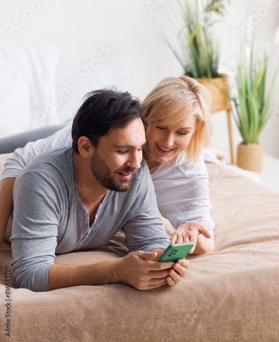 Playful couple lying in bed browsing internet read text message on modern cellphone cell phone cellular device. Happy man and woman have fun shopping online by smartphone. Planning pregnancy concept.