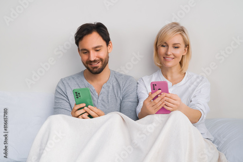 Couple lying in bed browsing internet read text message on modern cellphone cell phone cellular. Happy man and woman have fun shopping online by smartphone. Planning pregnancy concept.