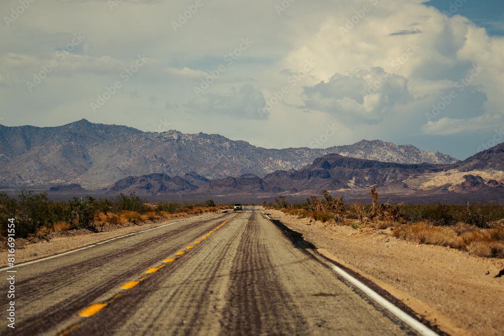 Car driving on a desert road against mountains, with a cloudy sky overhead in the United States Car driving on a desert road against mountains, with a cloudy sky overhead in the United States