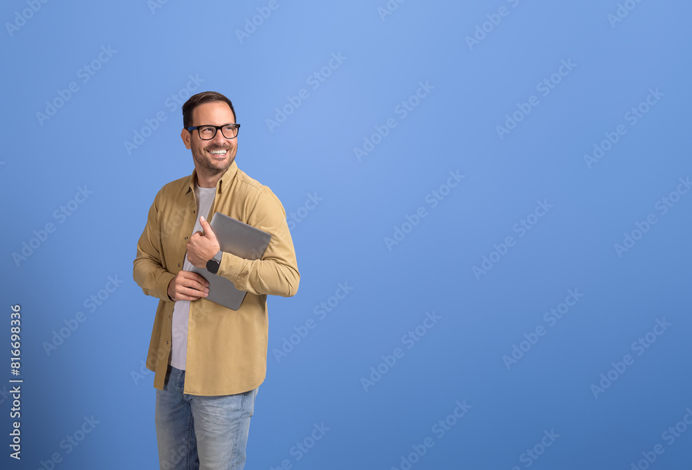 Portrait of positive young businessman holding laptop and looking away cheerfully on blue background Portrait of positive young businessman holding laptop and looking away cheerfully on blue background