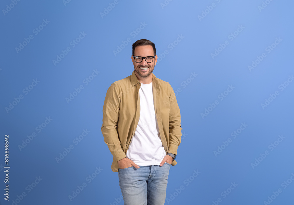 Portrait of handsome young man in eyeglasses and with hands in pockets standing on blue background