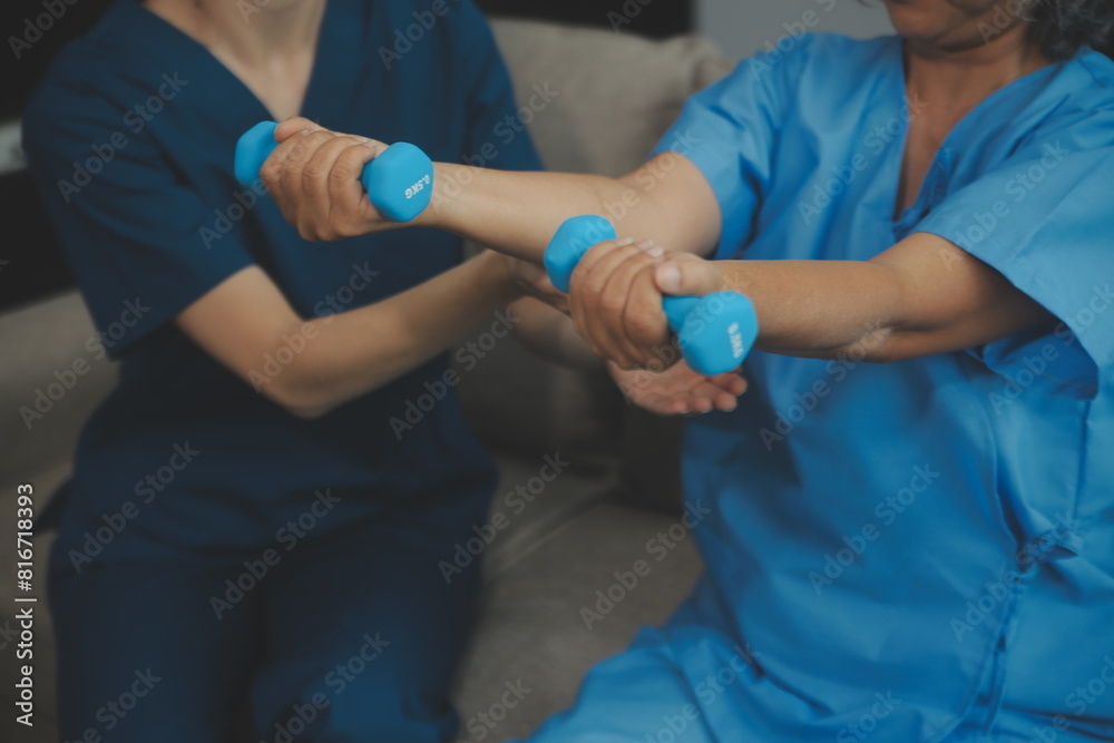 Old woman training with physiotherapist using dumbbells at home. Therapist assisting senior woman with exercises in nursing home. Elderly patient using dumbbells with outstretched arms.