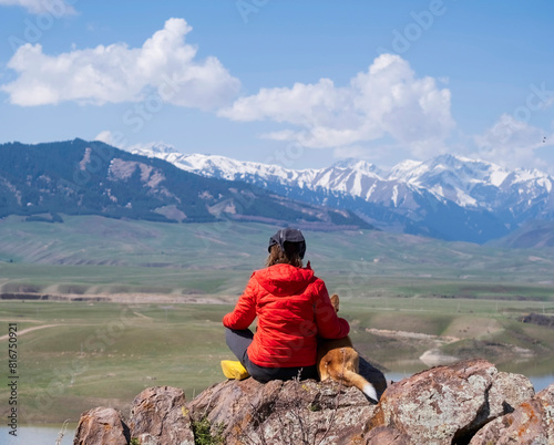A woman and a dog are sitting on a rock against the backdrop of a lake and a mountain with snow caps.