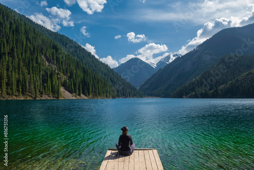 A woman tourist, admires the view from a wooden pier on a mountain lake with turquoise clear water. Lake Kolsai or Kulsay in the mountain range of Kazakhstan.