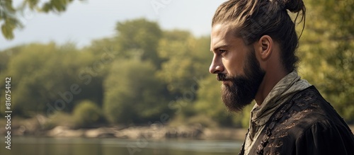 A bearded man with a stylish hair bun and make up is seen turned to the side looking away while speaking with a picturesque background of a tree and a river. Copyspace image photo