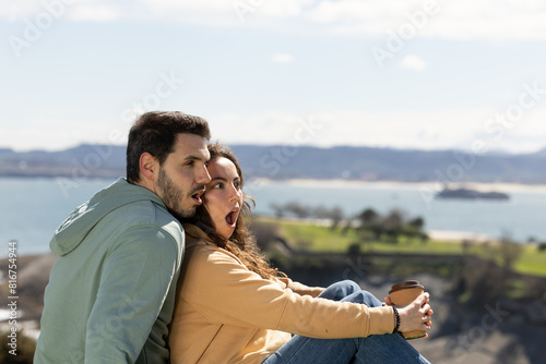 Surprised couple with the sea in the background. Couple at a viewpoint with the blue sea in the background while they are amazed at something they see. Surprised couple while holding a reusable glass.