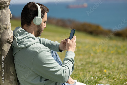 man listening to music with his green wireless headphones while choosing songs on his mobile phone. Listening to podcasts with headphones while checking your social networks, news or shopping online.