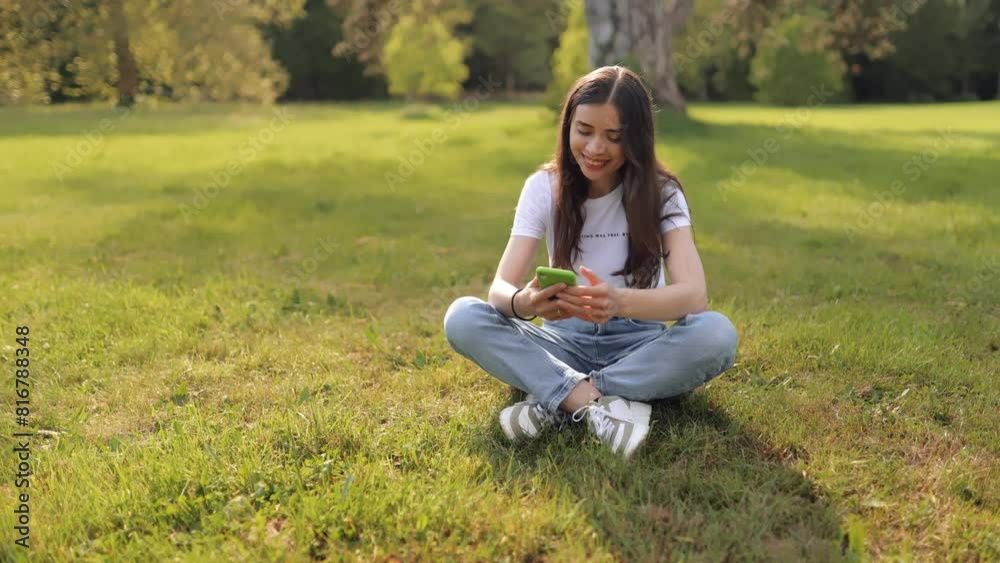 Young attractive woman girl student sitting on lawn in grass in park on lawn and using smartphone, smiling, shopping online, texting with friends or studying remotely Young attractive woman girl student sitting on lawn in grass in park on lawn and using smartphone, smiling, shopping online, texting with friends or studying remotely
