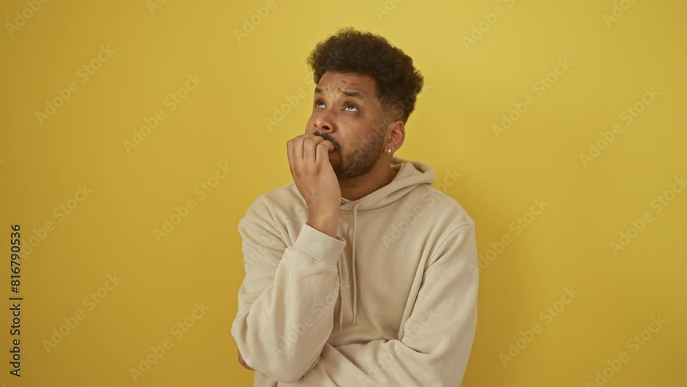 Anxious african american man in hoodie nervously biting fingernails, exhibiting stress, problem, fear over yellow isolated background. Anxious african american man in hoodie nervously biting fingernails, exhibiting stress, problem, fear over yellow isolated background.