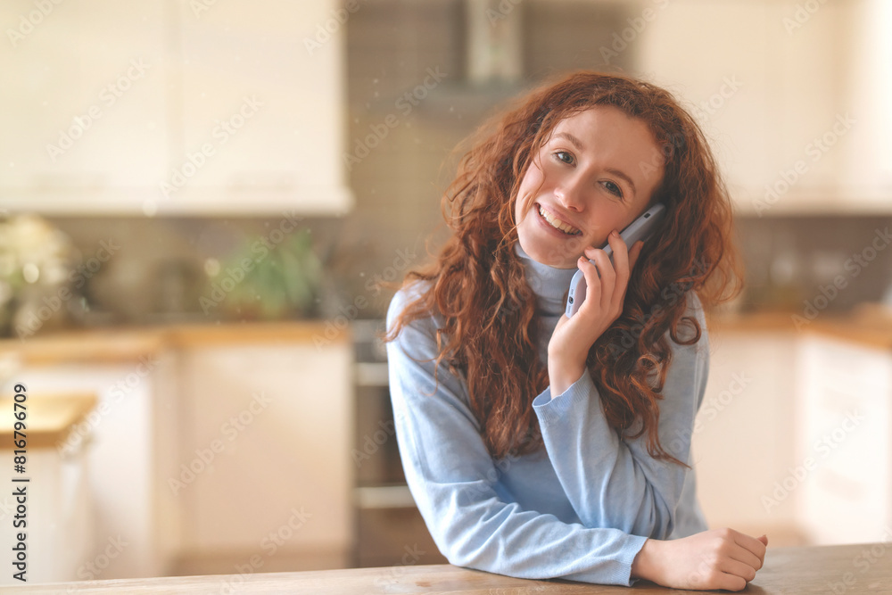 Fototapeta premium joyful woman using mobile phone, talking sitting in kitchen at home 