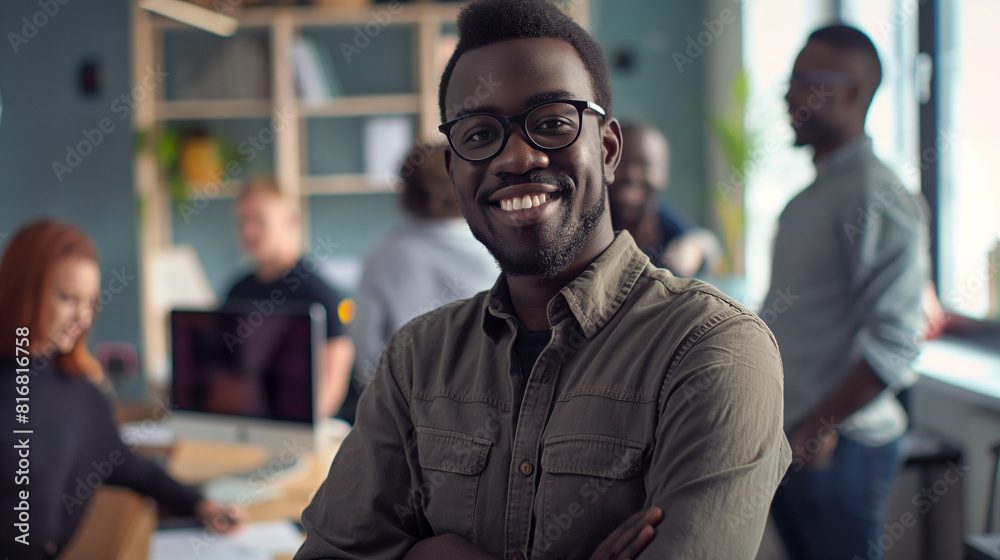 A confident, smiling multi-ethnic man stands in an office, facing the camera, with a diverse team working in the background. Reflects youthfulness and professionalism in a diverse business environment A confident, smiling multi-ethnic man stands in an office, facing the camera, with a diverse team working in the background. Reflects youthfulness and professionalism in a diverse business environment