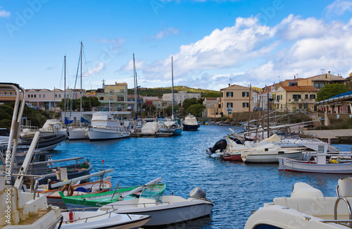 Vista del puerto de Stintino en Cerdeña, Italia, durante la primavera. La imagen muestra una variedad de barcos y yates atracados en el puerto, rodeados de edificios mediterráneos. 