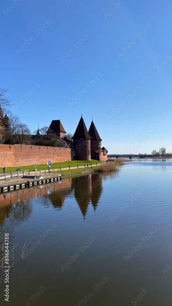 Medieval brick castle with conical towers and surrounding wall reflected in calm water under a clear blue sky on a sunny day