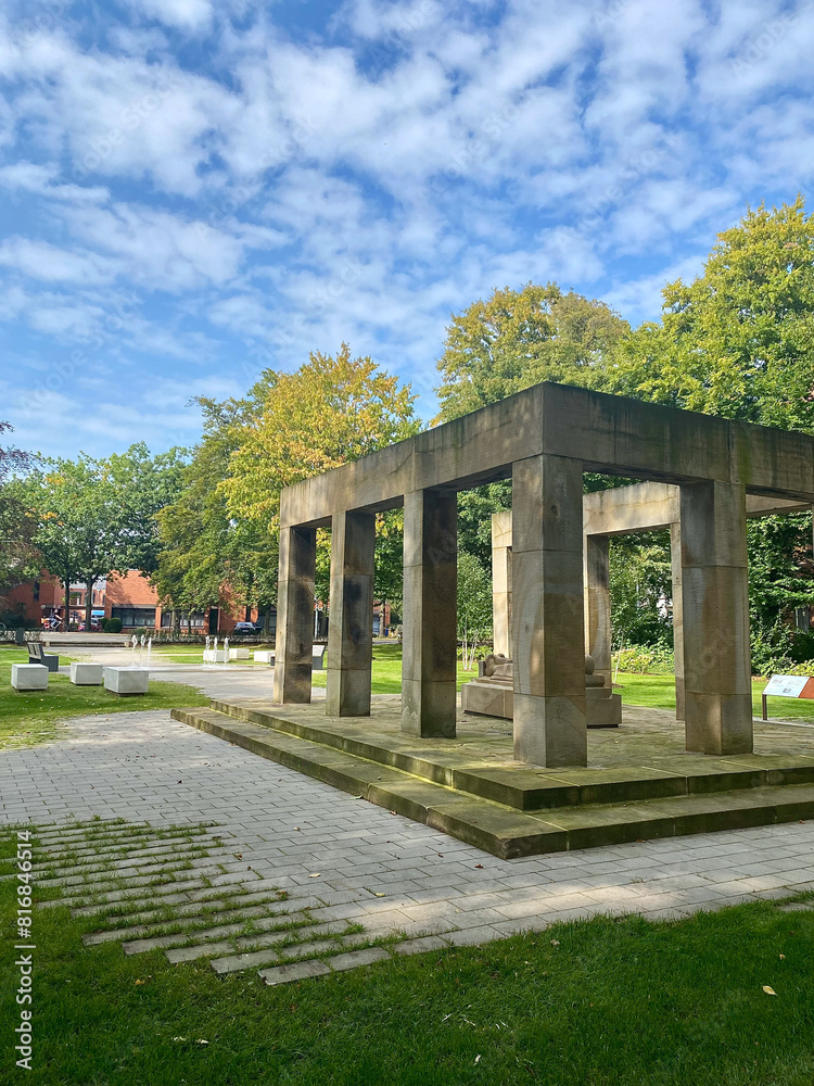 Stone monument in a green park with lush trees and a pathway under a partly cloudy sky. Tranquil setting with benches and manicured lawns