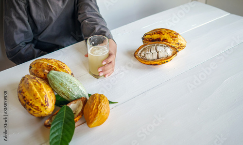 Fresh Cacao water in glass and half sliced ripe yellow cacao pod with white cocoa seed in the hands of boy teen, Cacao juice in glass on white wooden table, Healthy lifestyle