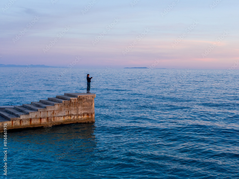 Naklejka premium Girl on a Pier Overlooking the Sea - Pink Sunset Sky