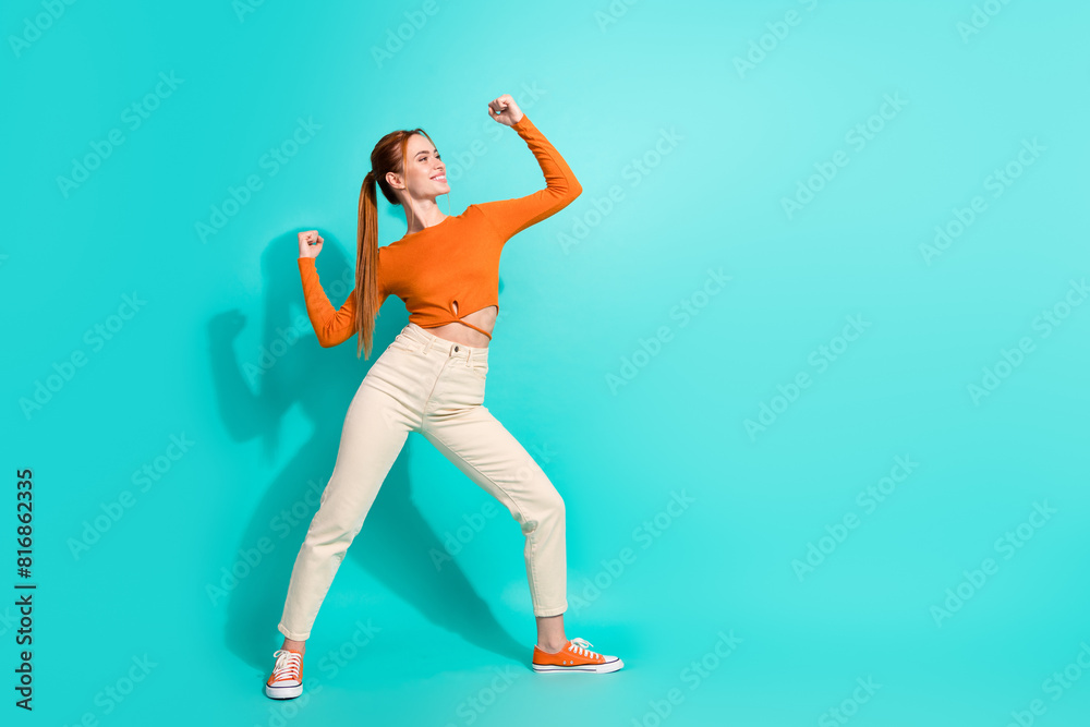 Full size photo of strong confident girl dressed orange shirt pants clenching fists look empty space isolated on teal color background Full size photo of strong confident girl dressed orange shirt pants clenching fists look empty space isolated on teal color background