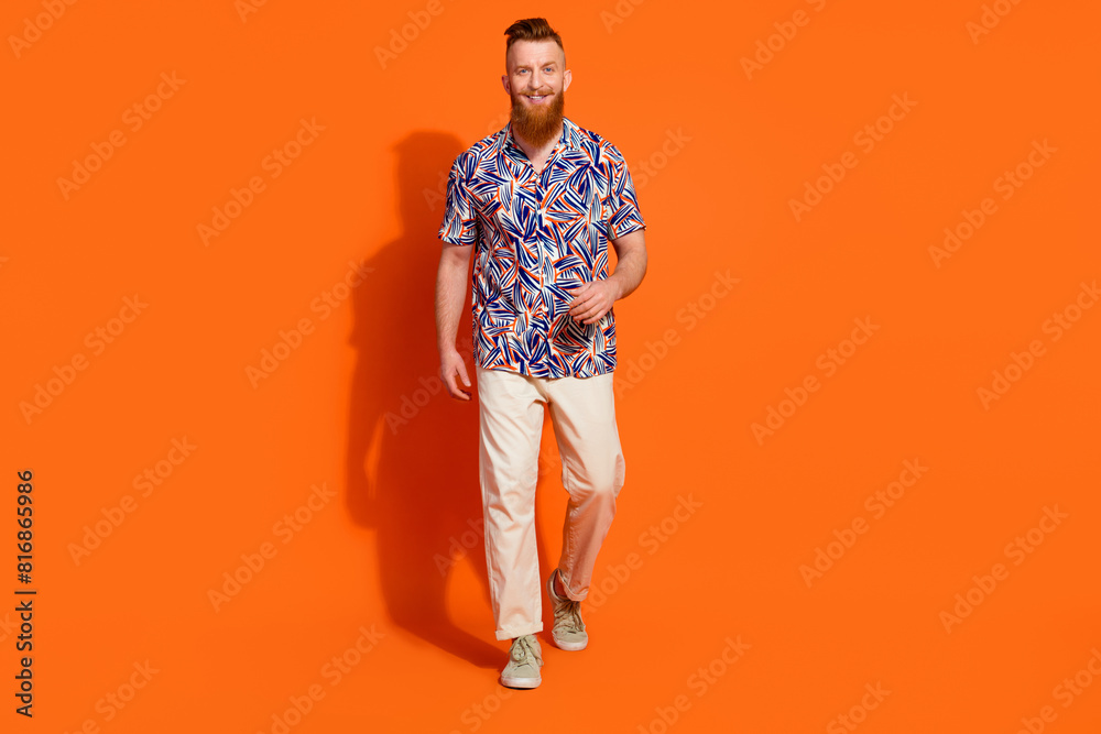 Full length photo of friendly handsome cool stylish guy wear print shirt white trousers walking isolated on orange color background Full length photo of friendly handsome cool stylish guy wear print shirt white trousers walking isolated on orange color background