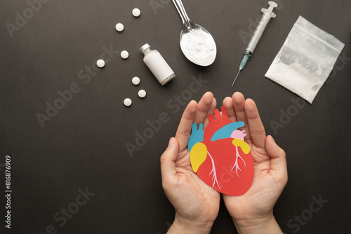 Effects of drugs on the human heart. Hands holding paper cut of heart with drug powder, syringe and pills on dark background. International Day against Drug Abuse.