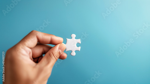 A close-up of a hand holding a single white puzzle piece against a plain blue background, symbolizing problem-solving, teamwork, and the last piece of the puzzle. photo
