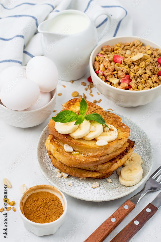 A stack of French toast, banana, nut, cinnamon and mint toast, a bowl of granola, milk and eggs. European breakfast on a light background, top view. A stack of French toast, banana, nut, cinnamon and mint toast, a bowl of granola, milk and eggs. European breakfast on a light background, top view.