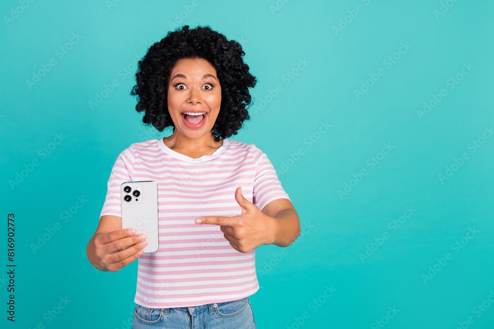 Portrait of young happy girlfriend in striped t shirt directing finger new release smart phone isolated on aquamarine color background Portrait of young happy girlfriend in striped t shirt directing finger new release smart phone isolated on aquamarine color background