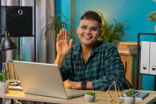 Hello. Indian man smiling friendly at camera and waving hands gesturing hello, hi, greeting or goodbye welcoming, online video call POV conversation. Guy at home office table workplace. Lifestyle