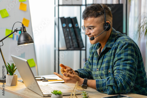 Hindu business man wearing headset freelance worker call center or support service operator helpline talking with client or colleague communication support. Guy sitting at home office desk. Lifestyle