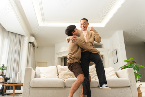 Asian middle aged son holding senior father, smiling, laughing, funny moment. Healthy son with strong bones and joints carrying old man in living room at home. Happy two generations of men in family