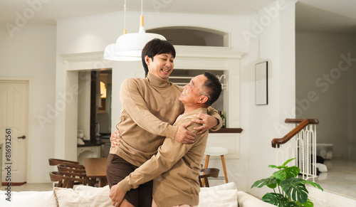 Asian senior father holding middle aged son, smiling, laughing, funny moment. Healthy dad with strong bones and joints carrying young man in living room at home. Happy two generations of men in family