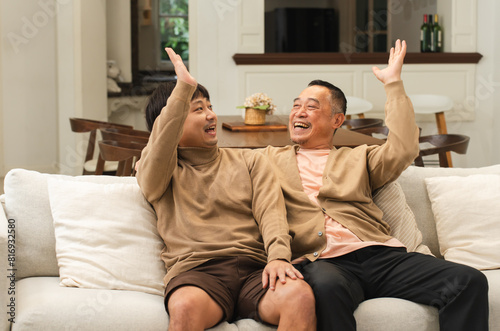 Cheerful Asian old man giving high five to happy middle aged son, celebrating good news. Smiling two male generations family feeling excited together, resting on sofa in living room at home